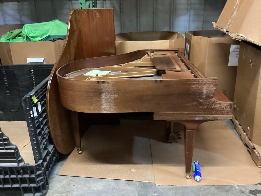 A distressed wooden grand piano is partially disassembled in a cluttered storage area.