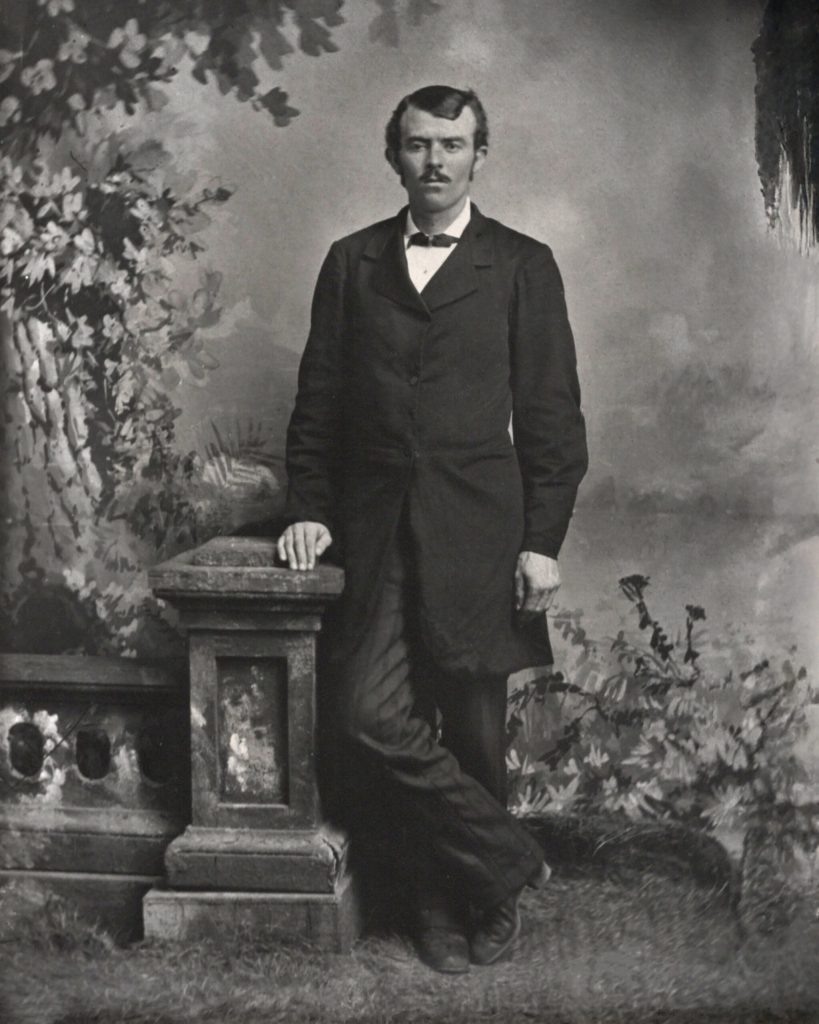 A vintage black and white portrait shows a man in a suit and bow tie leaning on a
