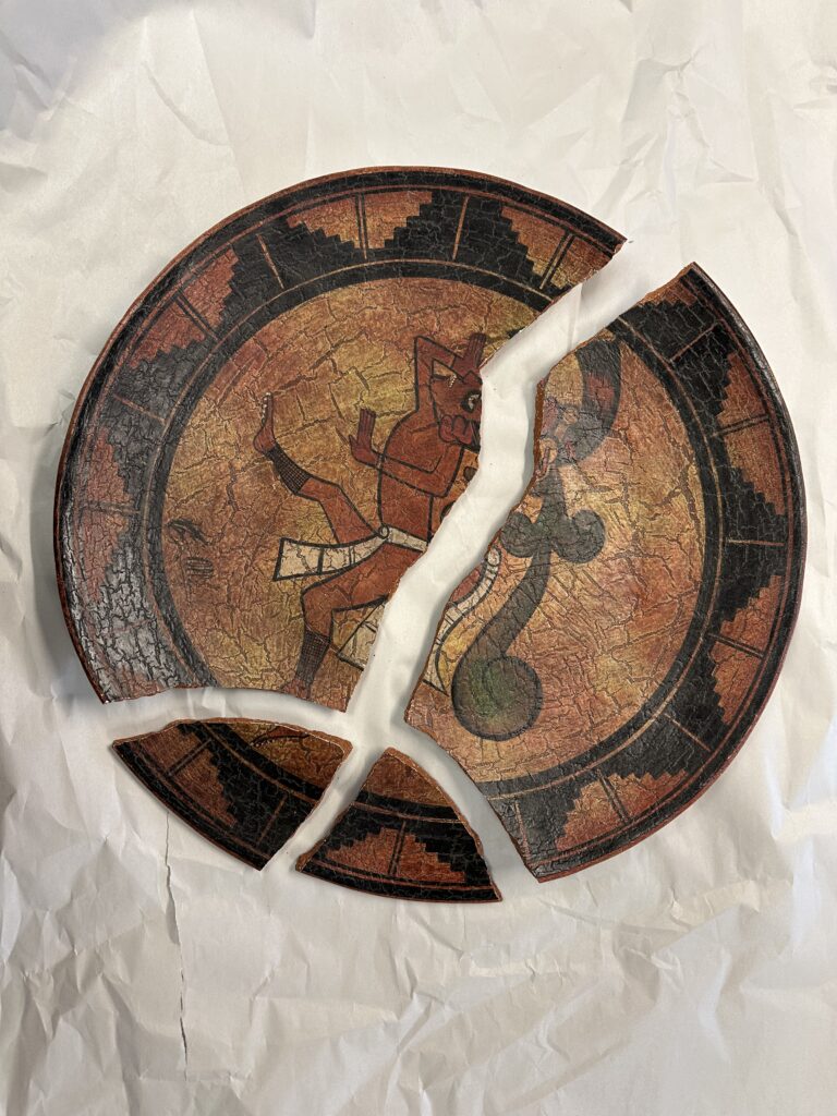 A broken Mesoamerican-style plate with a red figure and serpent design lies on