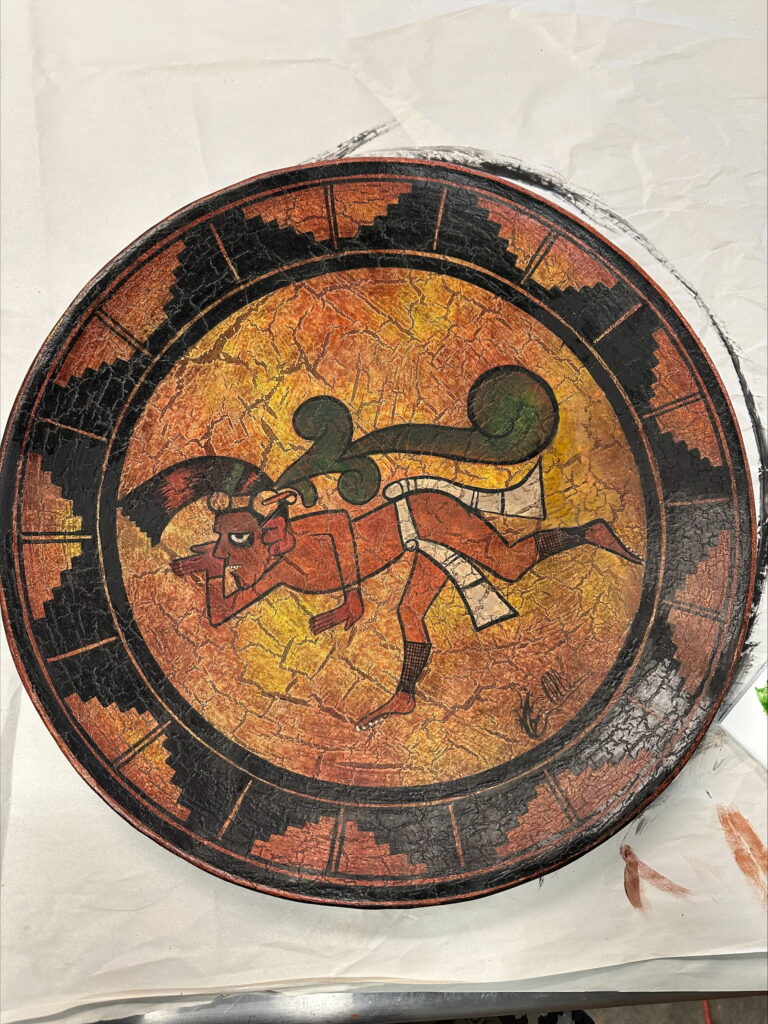 A round, textured decorative plate depicts a red, stylized Mesoamerican figure wearing a he