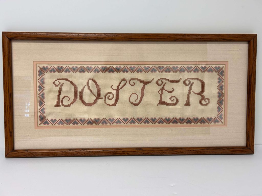 Framed cross-stitch art spells "DOSTER" in brown, ornate letters with patterned pink and blue borders.