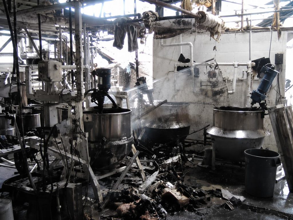 Inside a fire-ravaged factory, large industrial vats and machinery are covered in soot and