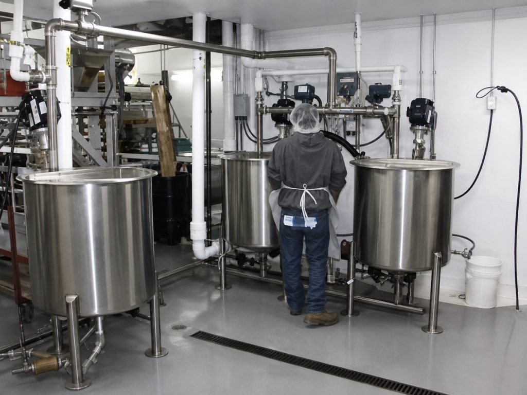 A worker in a hairnet and apron stands among stainless steel processing tanks in a clean industrial