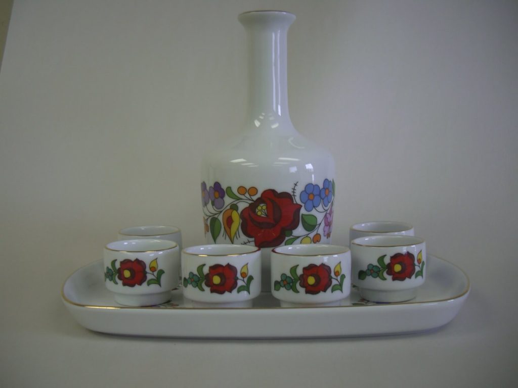 A white ceramic decanter and six small cups with colorful folk art floral designs sit on