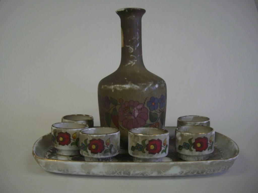 A vintage floral pottery decanter and six matching small cups sit on an oval tray. TITLE