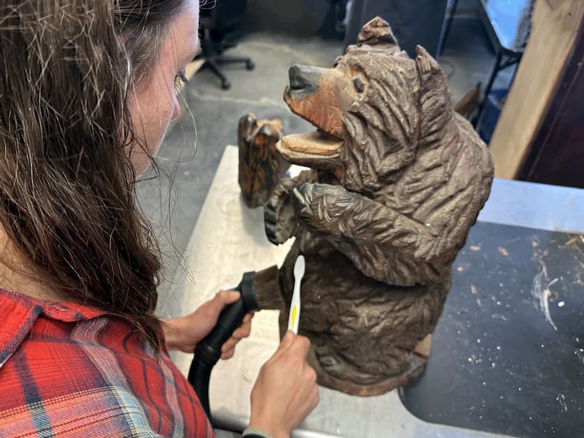 A woman in a plaid shirt carefully cleans a wooden bear carving with a vacuum and
