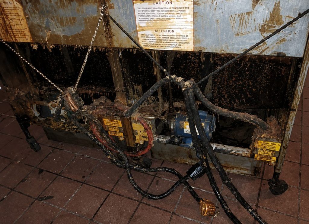 Grime-covered industrial equipment with exposed electrical components and chains on a tiled floor. TITLE