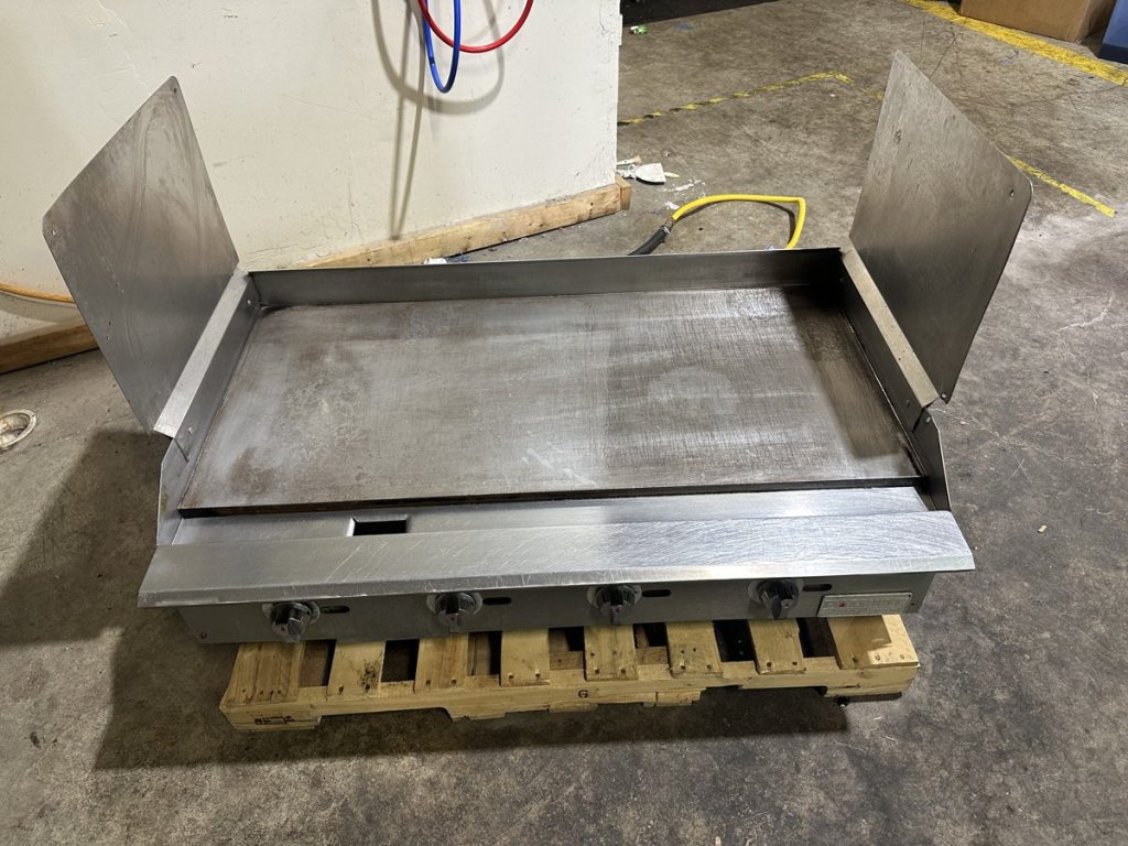 A used commercial stainless steel griddle with three control knobs rests on a wooden pallet. TITLE