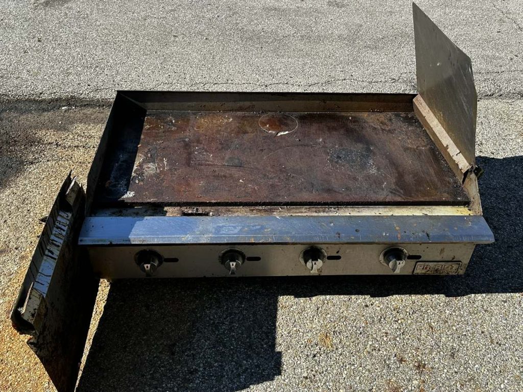 A dirty, used commercial flat-top griddle with three control knobs sits outdoors on