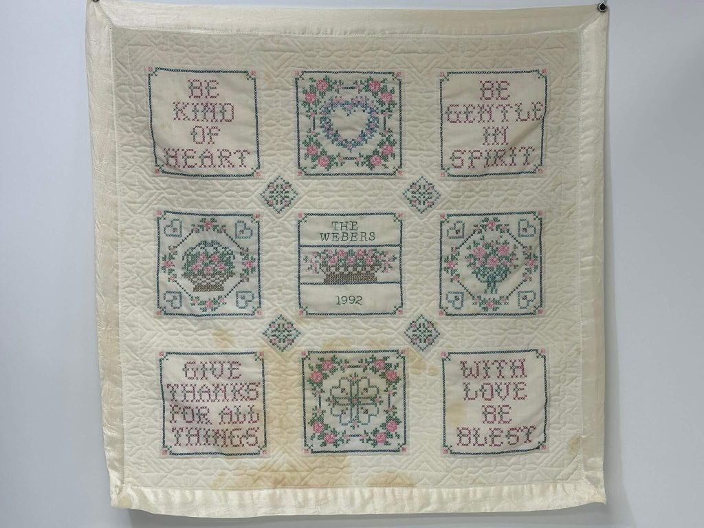 A cream quilted wall hanging features cross-stitched inspirational phrases, floral designs, and "THE WEBERS