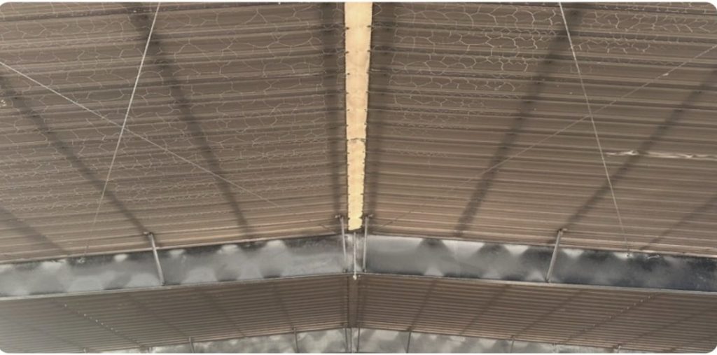 Corrugated metal roof interior showing a cracked surface texture, support beams, and wires. TITLE