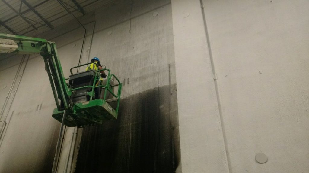 A worker in a boom lift pressure washes a large industrial wall, revealing a clean, light