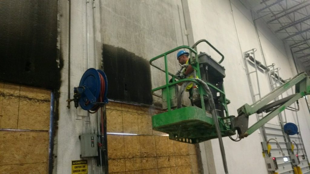A worker on a green scissor lift is pressure washing a dirty industrial wall.