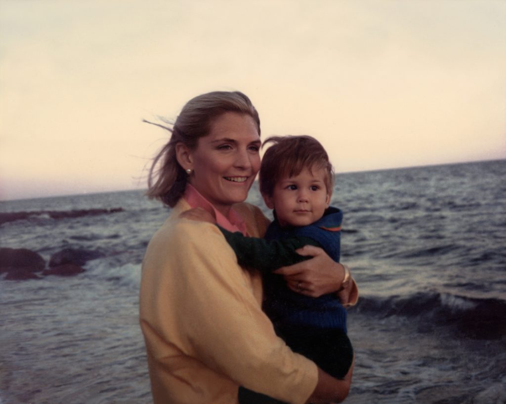 A smiling woman holds a young child at the windy seaside with the ocean behind them.
