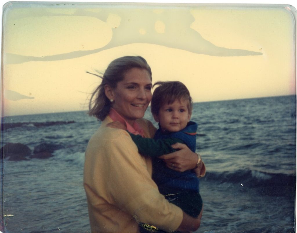 A smiling woman holds a young child by the ocean under a light sky.