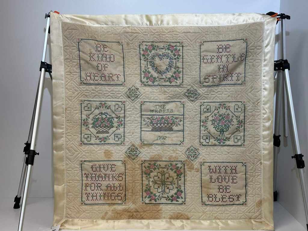 A cream-colored cross-stitched quilt from 1992 with inspirational phrases and