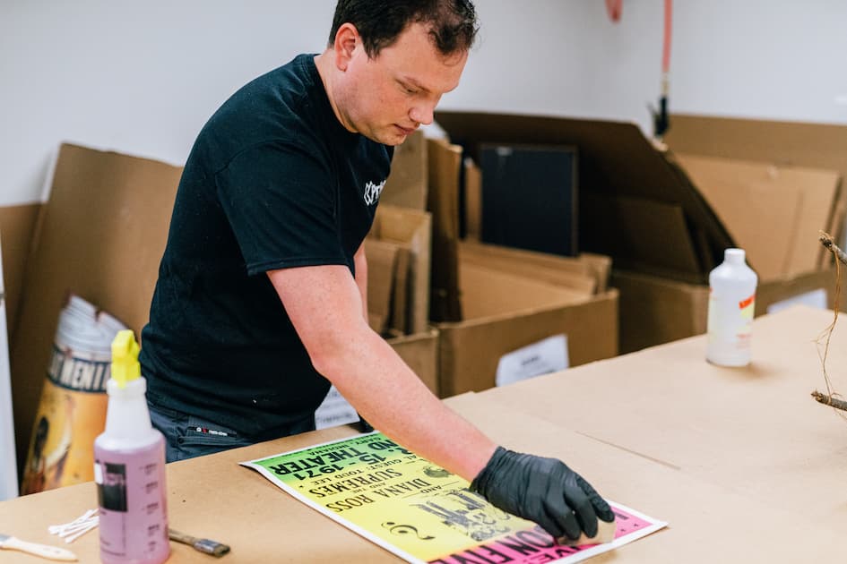 A person in black gloves carefully works on a vibrant vintage concert poster on a brown table.