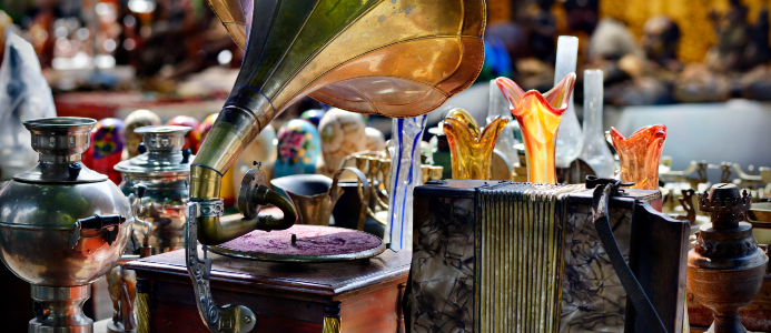 Table full of antiques such as a gramophone, accordion, colored glass and metal vases, and more.