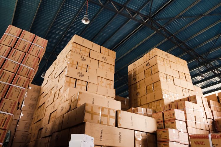 Tall stacks of cardboard boxes fill a warehouse under a blue corrugated metal ceiling. TITLE