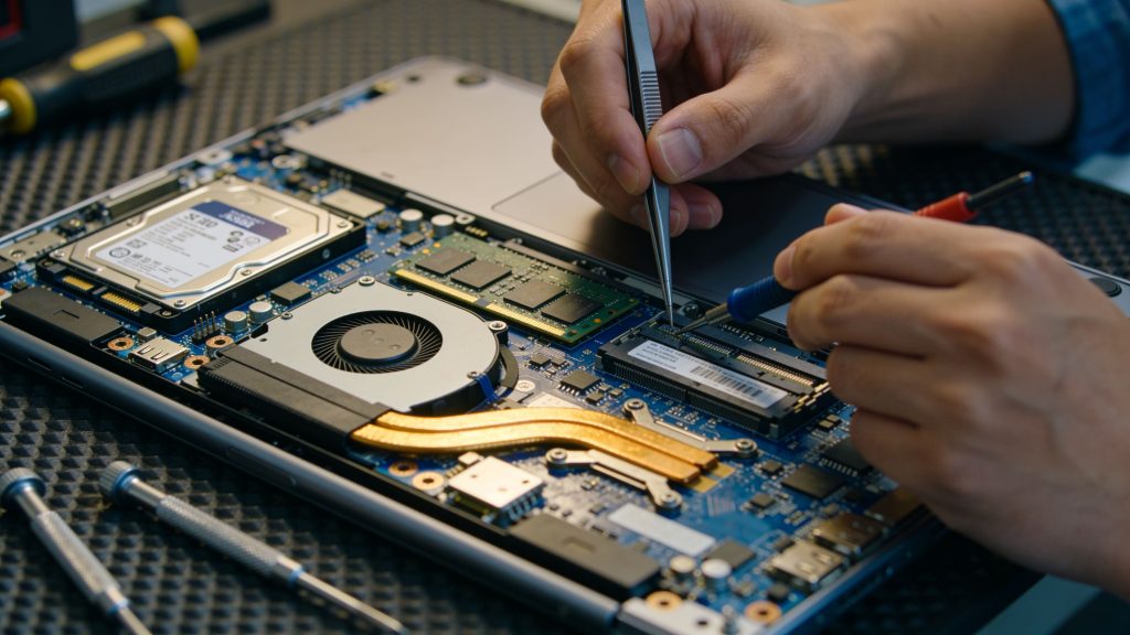 A person's hands use precision tools to repair the open internal components of a laptop.