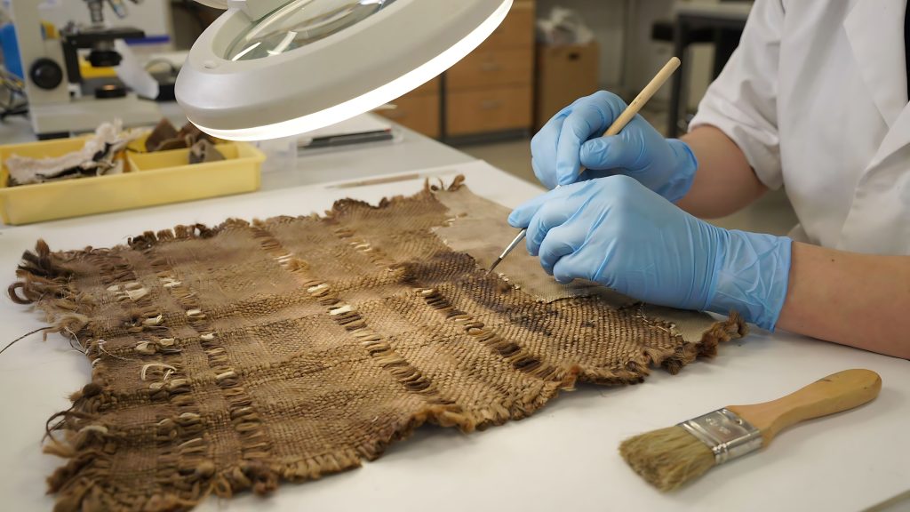 Gloved hands carefully clean an ancient, woven brown textile under a bright magnifying lamp.