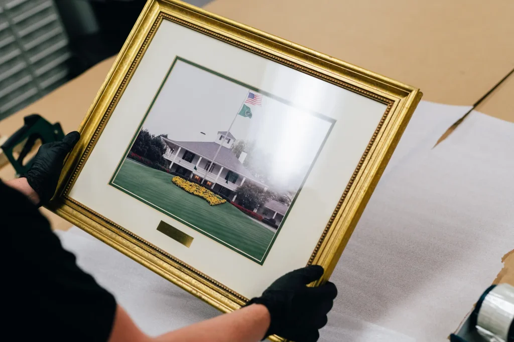 Gloved hands hold a framed print of the iconic Augusta National Golf Club clubhouse.