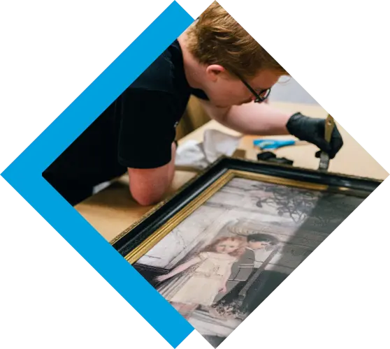 A person in glasses and black gloves carefully cleans a framed vintage artwork of two children. TITLE
