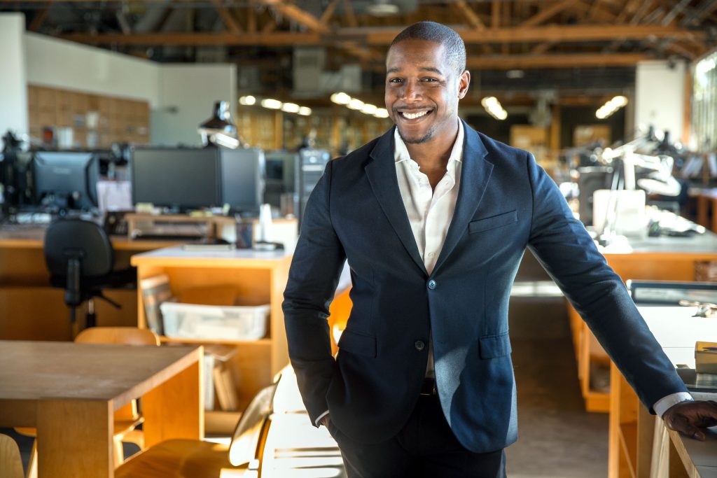 A happy Black businessman in a suit jacket poses in a bright, open-plan office.