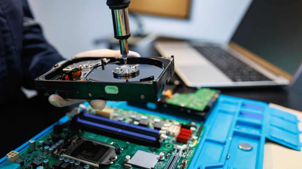 A person uses a soldering iron to work on an open hard drive, surrounded by computer components.