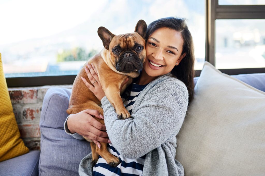 A smiling woman hugs her fawn French Bulldog while sitting on a couch by a window.