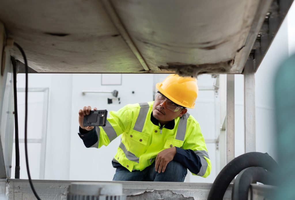 A technician in a hard hat and safety vest photographs industrial equipment with a smartphone. TITLE: