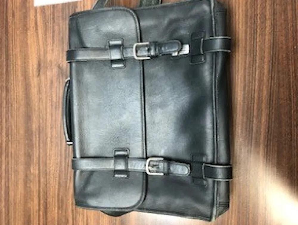 A black leather briefcase with two buckled straps lies on a wooden surface. TITLE: