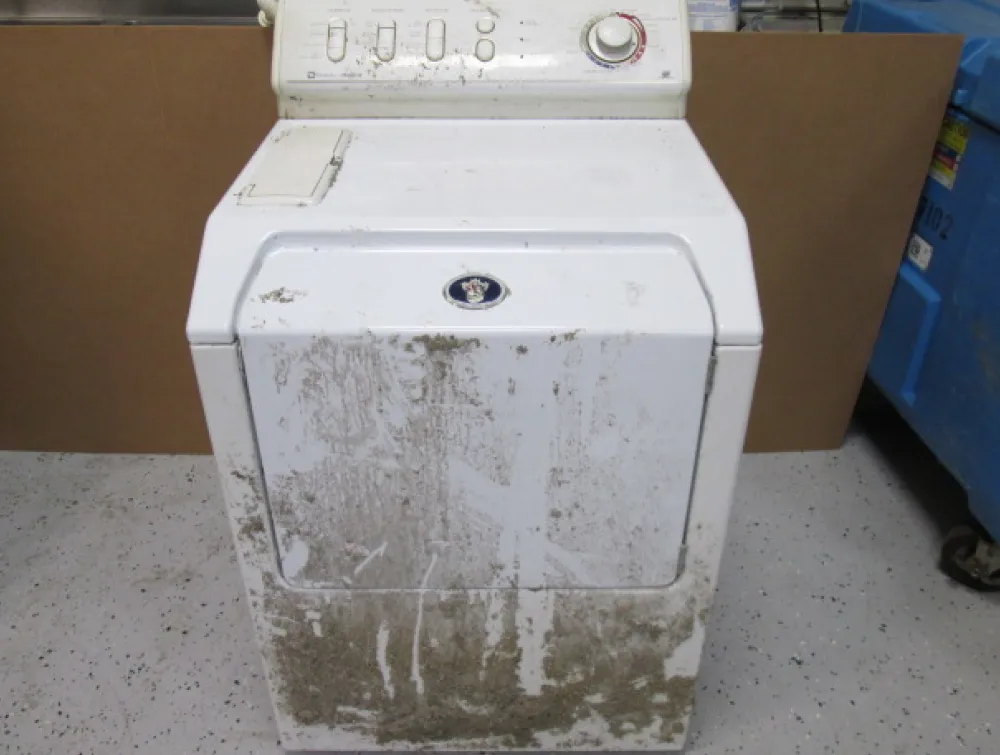 A dirty white top-load washing machine is caked with mud and grime on its front